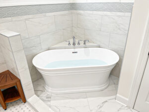 White freestanding soaking bathtub with chrome fixtures, marble-look tile surround, and decorative gray mosaic tile border.